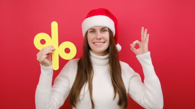 Merry pretty fun young woman wear warm white sweater and Xmas Santa hat holding big paper yellow percent mark, posing isolated over pastel red studio background. Happy New Year 2024 holiday concept