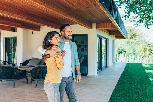 Couple standing in front of their new home. They are both wearing casual clothes and embracing. They are looking away and smiling. The house is contemporary with porchway and a green lawn, wood and glass exterior design