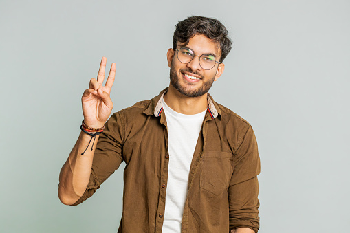 Happy Indian young man showing victory sign, hoping for success and win, doing peace gesture, smiling with kind optimistic expression, celebrate. Arabian Hindu guy isolated on gray studio background