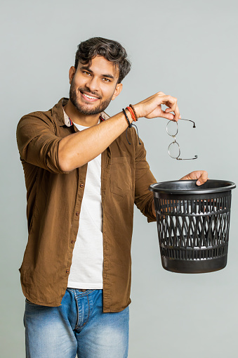 Indian man taking off throwing out glasses into bin after medical vision laser treatment therapy surgery looking smiling at camera, heal, cure. Arabian guy isolated on studio gray background. Vertical