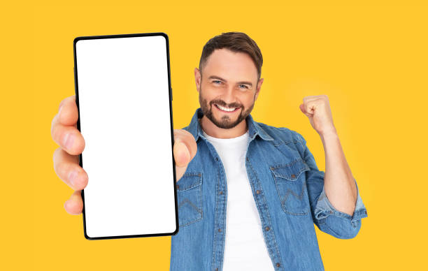 istock Handsome joyful man holding a Smartphone mockup with a blank screen and smiling on an orange background. 1735643351