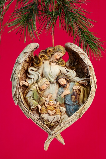 Christmas nativity ornament of the Holy Family surrounded with angel wings, hanging from Christmas tree branch, on red background. Vertical image would be good for Christian or religious Christmas use.