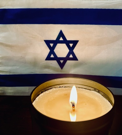 A lit candle in a black metal tin illuminates an Israeli flag in a Hoboken, New Jersey apartment. The National Flag of the State of Israel features a blue hexagram (symbolizing the Star of David) on a white background centered between two blue stripes. It was adopted on October 28, 1948.