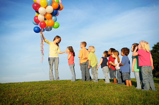 Color photo of children waiting in line to get balloons from a teenage girl.