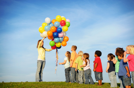 Color photo of a group of children waiting in line to get balloons from a teenage girl.