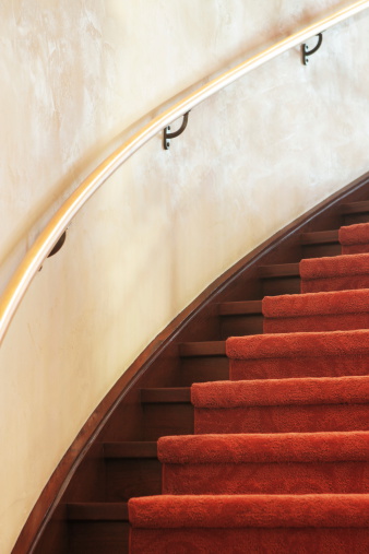 Curved spiral staircase with red carpet and wood handrail on stucco wall.