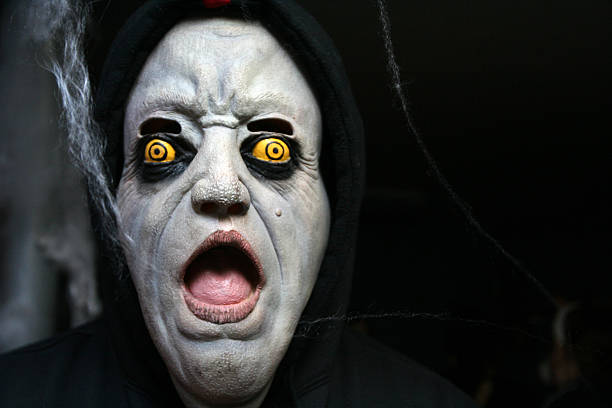 Scarey halloween mask with black copyspace to the right. Mask is white with a furrowed brow. Eyes are bright yellow encircled in black. Mouth is wide open with wearer's tongue showing through. Spider webish material hangs down. A black hood completes the costume.