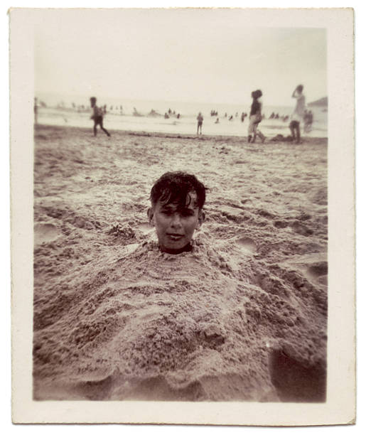 Vintage photo of boy buried in sand at the beach Head poking out from the sand.  Image taken in 1957.  Note that the image has soft focus and grain and grunge because of its age sepia toned photos stock pictures, royalty-free photos & images