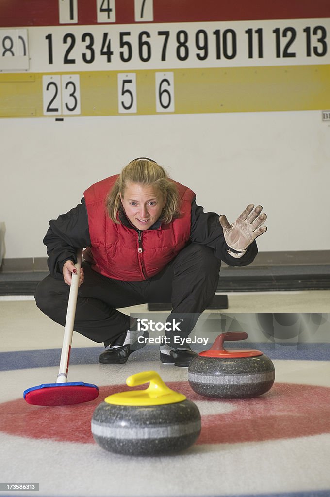 Calling the Shot in Curling Game Curling game. Focus on curler checking position of stones (rocks) in the house which is the target. She squats with her curling broom and holds up hand. Curling is an official . winter sport. Curling - Sport Stock Photo