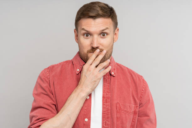 istock Amazed confused man with raised eyebrows covering mouth with hand looking at camera on studio wall 1735864487