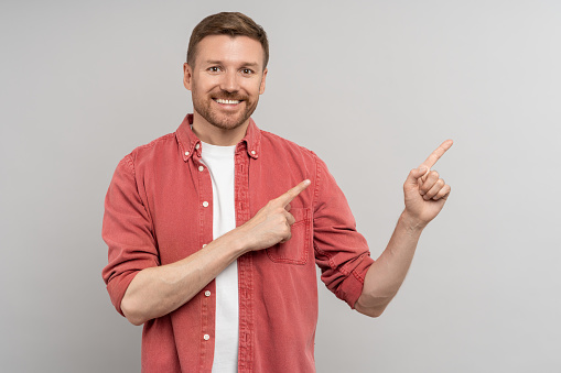 Excited happy positive young guy student pointing aside with fingers hand gesture at copy space advertising product, presenting sale discount promo offer standing isolated on gray background