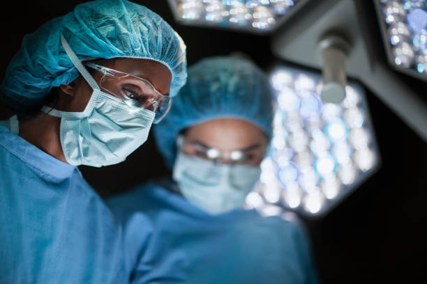 Asian Indian ethnicity medical professional doctor and nurse looking down to patient in surgery operation room in hospital with lights stock photo