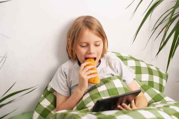 istock A boy sits on the bed, absorbed in his tablet, leisurely enjoying a bun 1735866026