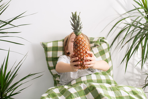 Lounging in bed, a 9-10-year-old boy with flowing locks leisurely holds a pineapple, embodying relaxation and the concept of doing nothing