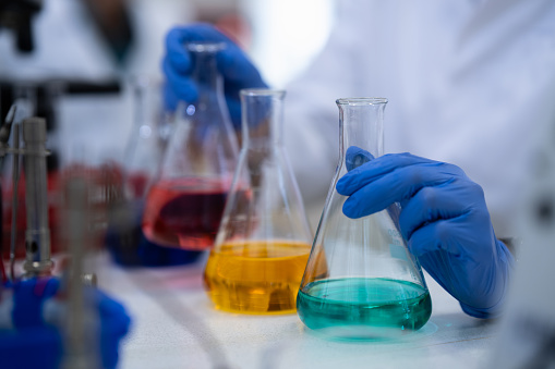 A research chemist holds three different chemicals in glassware on a table with his gloved hands.
