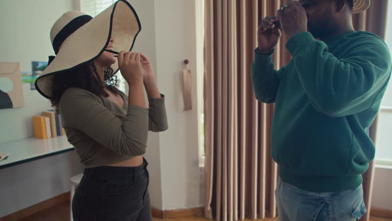 Cheerful Multiethnic Couple Trying on Sun Hats and Sunglasses at Home