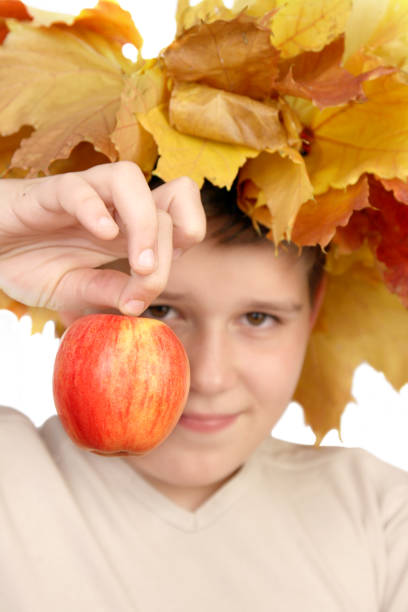 Vertical 4-year-old cute Caucasian boy with wreath of red maple leaves on his head against background of autumn nature meadow,making various signs with his fingers.Selective focus, blurred background.