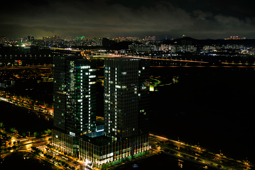 Incheon City Skyline at night in South Korea, tall clusters of illuminated buildings on the dark cloudy sky with the view of Seoul over the distant mountains