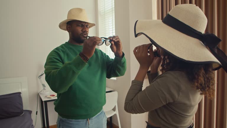 Young Couple Trying on Straw Hats and Sunglasses while Packing for Honeymoon