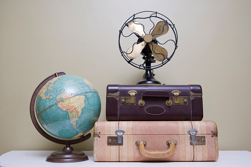 Selection of various vintage, antique and retro items including two suitcases, a globe and a fan that sit on a white table 