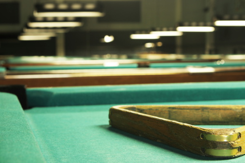 Pool hall lighting on pool table felt surface with wooden rack and depths of billiard room stretching into darkness beyond.