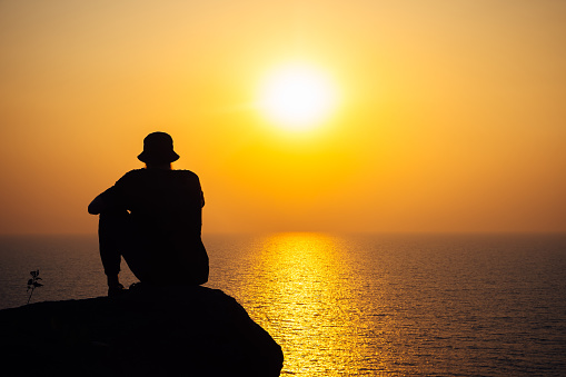 The silhouette of a man in a hat against the background of a bright sunset and the endless sea. A thoughtful adventurer, thoughts about the future and the meaning of life.