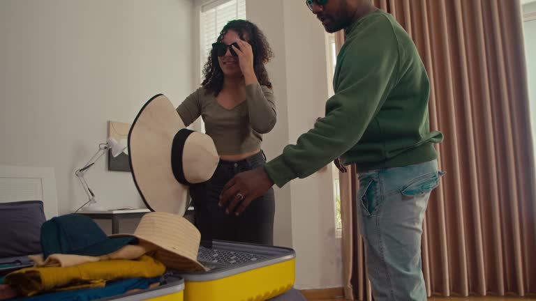 Happy Husband and Wife Trying on Sun Hats and Sunglasses while Packing Suitcase