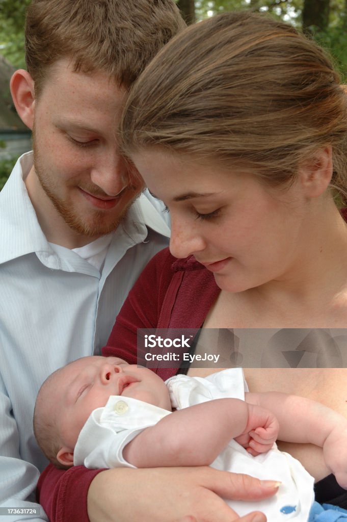 Young New Family A young mother and father dream of beginnings and the future with their new baby. Image is closely cropped to show faces of father, mother and infant. Father’s face is partially hidden. Mother holds baby and both parents look down at sleeping child with smiles on their faces. Adult Stock Photo