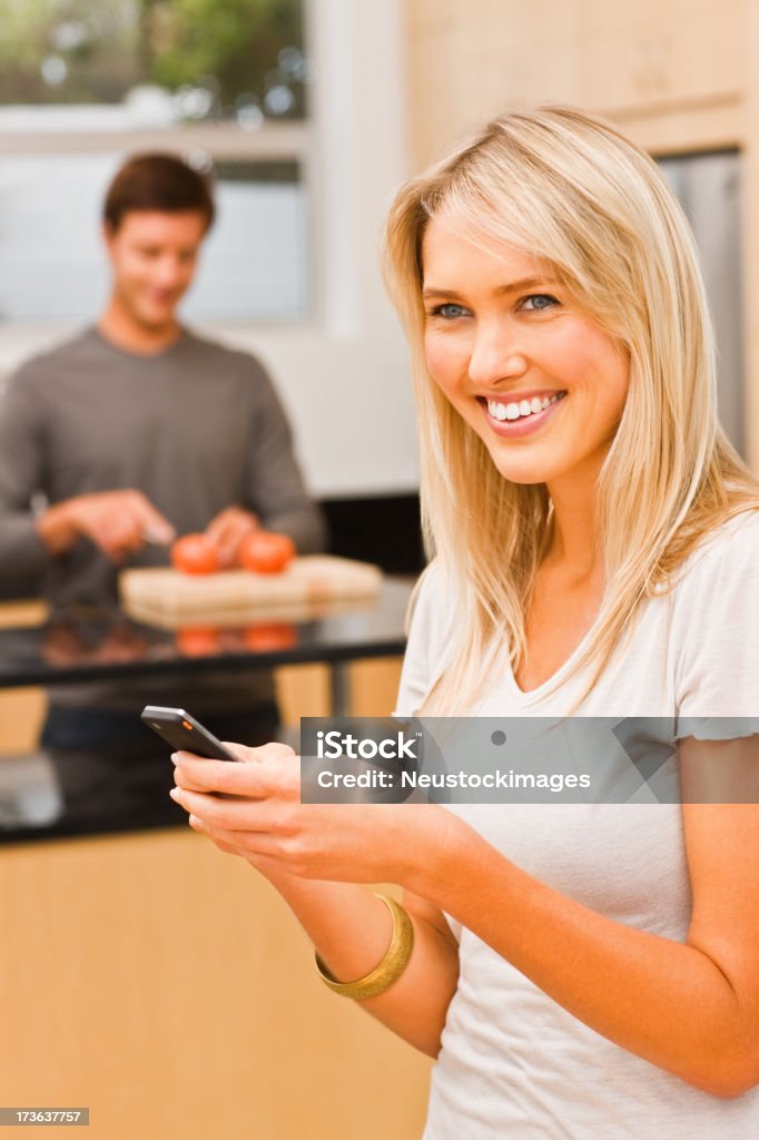 Giovane donna con cellulare mentre uomo taglio verdure in background - Foto stock royalty-free di Abbigliamento casual