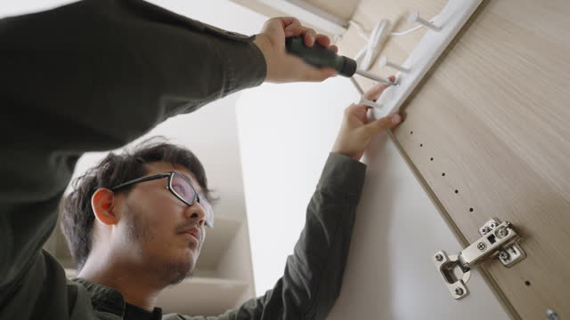Low angle and tilt up shot of a man opening closet door tightening screws on the hinge using screwdriver during a DIY flat-pack furniture assembly at home.