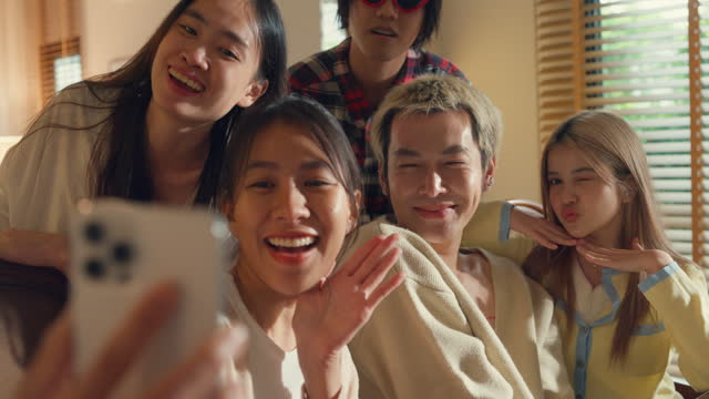 Closeup group of young Asian people using mobile phone taking selfie and having fun sitting at dining table at home. Multicultural friends enjoying spending together college house party.