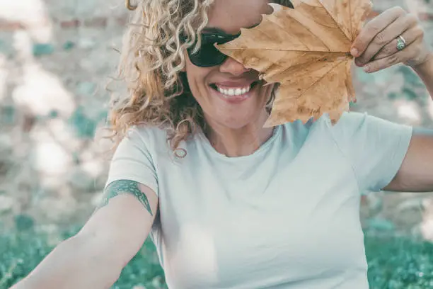 Playful happy woman portrait smiling and hidden half face with big autumn dry yellow maple leaf. Cheerful young adult female people smile and lok on camera wearing sunglasses in outdoor leisure day