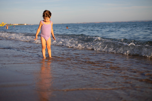 A three-year-old girl stands on the seashore and looks at her feet covered in seawater. The toddler is playing on the beach,