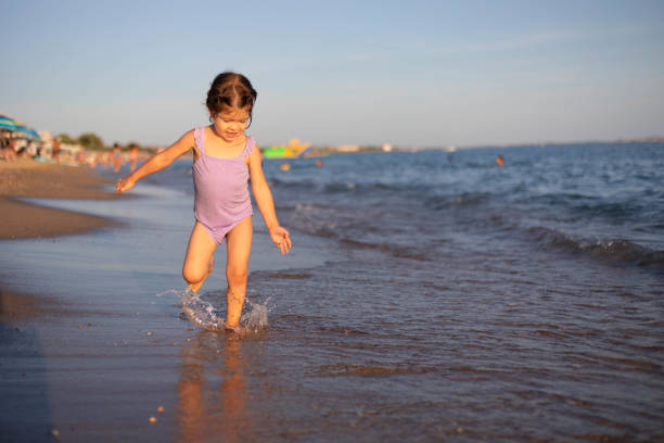 A child runs happily along the seashore and enjoys splashing with seawater. The three-year-old girl in the purple swimsuit is playing on the beach.
