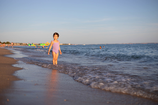 The adorable three-year-old girl is standing on the seashore. The portrait of a toddler standing and playing on the beach. The child is waiting for the sea wave to wash over her feet.