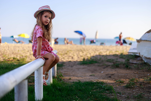 A child is sitting on a wooden pole and resting. There is a white pole (fence) on the beach, and the girl uses it as a chair.
