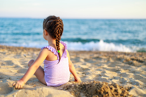 The little girl sits on the beach after swimming and looks at the sea. A child plays with sand while resting on the seashore.