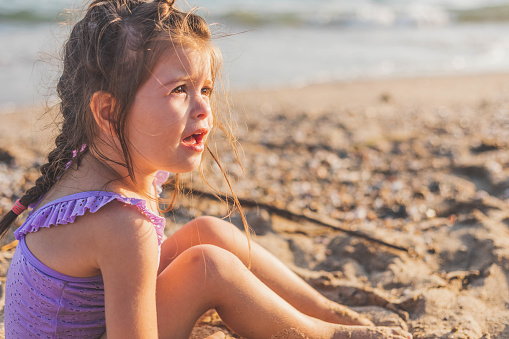 The little girl is sitting on the beach and crying. The children can't get everything, so, sometimes they just need to cry for no reason. The cute, little girl is showing her emotion, by sitting on the ground and crying.