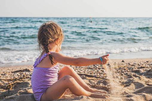 The cute, little girl is sitting on the beach and looking at the sea while playing with the sand. The child is trying to catch the sand with her hands.
