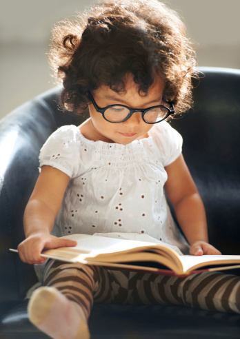 A cute little girl wearing glasses reading a book on the couch