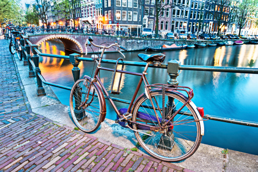 Typical Amsterdam City Scene at night. Visible are one of the many beautiful bridges, one bike, traditional dutch houses in raw and many street light and their reflection in the background. Amsterdam, Netherlands.