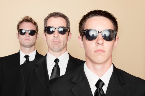 This is a horizontal, color image of three men of various ages dressed in black suits and ties. Sunglasses cover their eyes as they stand staggered with deadpan expressions.