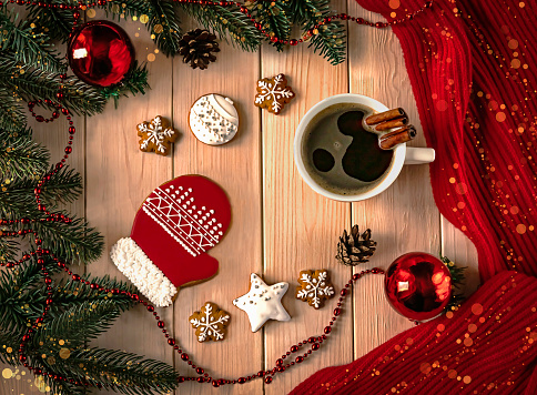 Christmas light wooden background with red gingerbread in the form of a glove and a cup of coffee with bokeh effect. Christmas tree balls, branches, necklace, cones and red scarf as decoration.