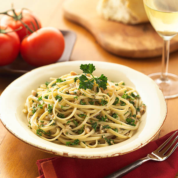 Linguini with white clam sauce with chopped parsley and garlic and a parsley sprig.  Shot with shallow focus on pasta with tomatoes, white wine and bread in the background.