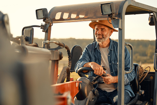 Mature handsome male farmer sitting in a tractor on a field on a hot day working on a farm. Farming and agriculture concept.