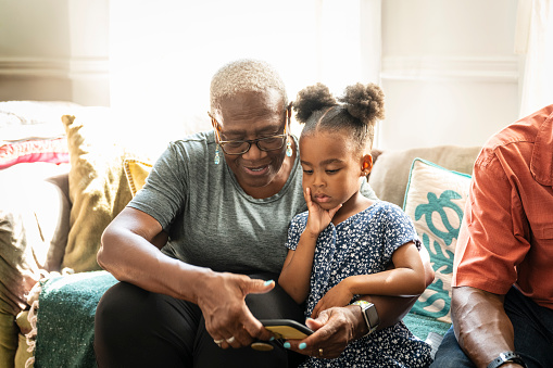 Black grandmother and granddaughter, 68 and 4 years of age, looking at portable device in living room of family home.