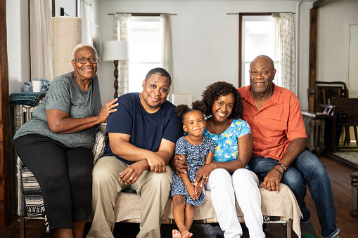 Front view of senior couple in their 60s, mature couple in 40s and 50s, 4 year old child, all together on sofa, looking at camera and smiling.