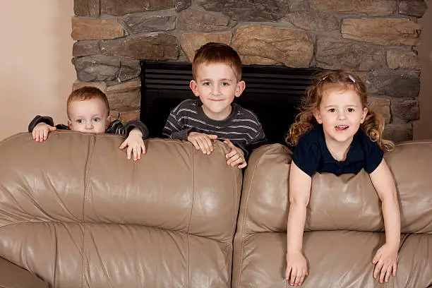 Three cute siblings looking over the back of a sofa.