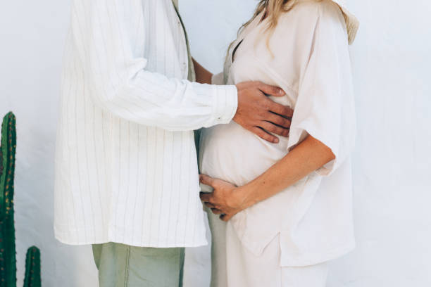 Unrecognizable pregnant couple touching belly while standing against white wall. Unrecognizable pregnant couple touching belly while standing against white wall. trophy wife stock pictures, royalty-free photos & images
