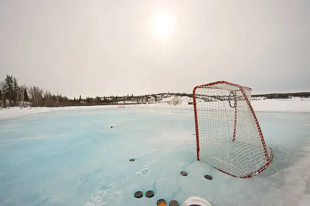 A weak sun shines through light overcast on an outdoor hockey rink in Canada's Arctic near Yellowknife. Pale blue ice with snow at the edges, net and pucks.  Grey sky for copy space.